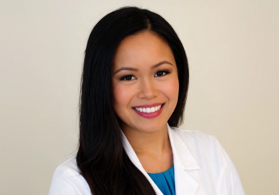 A horizontal, mid-shot portrait of a smiling woman with long, straight dark brown hair. She is wearing a white professional medical lab coat over a vibrant teal pleated top, posed against a solid off-white background. The name embroidery on the coat is cropped out.